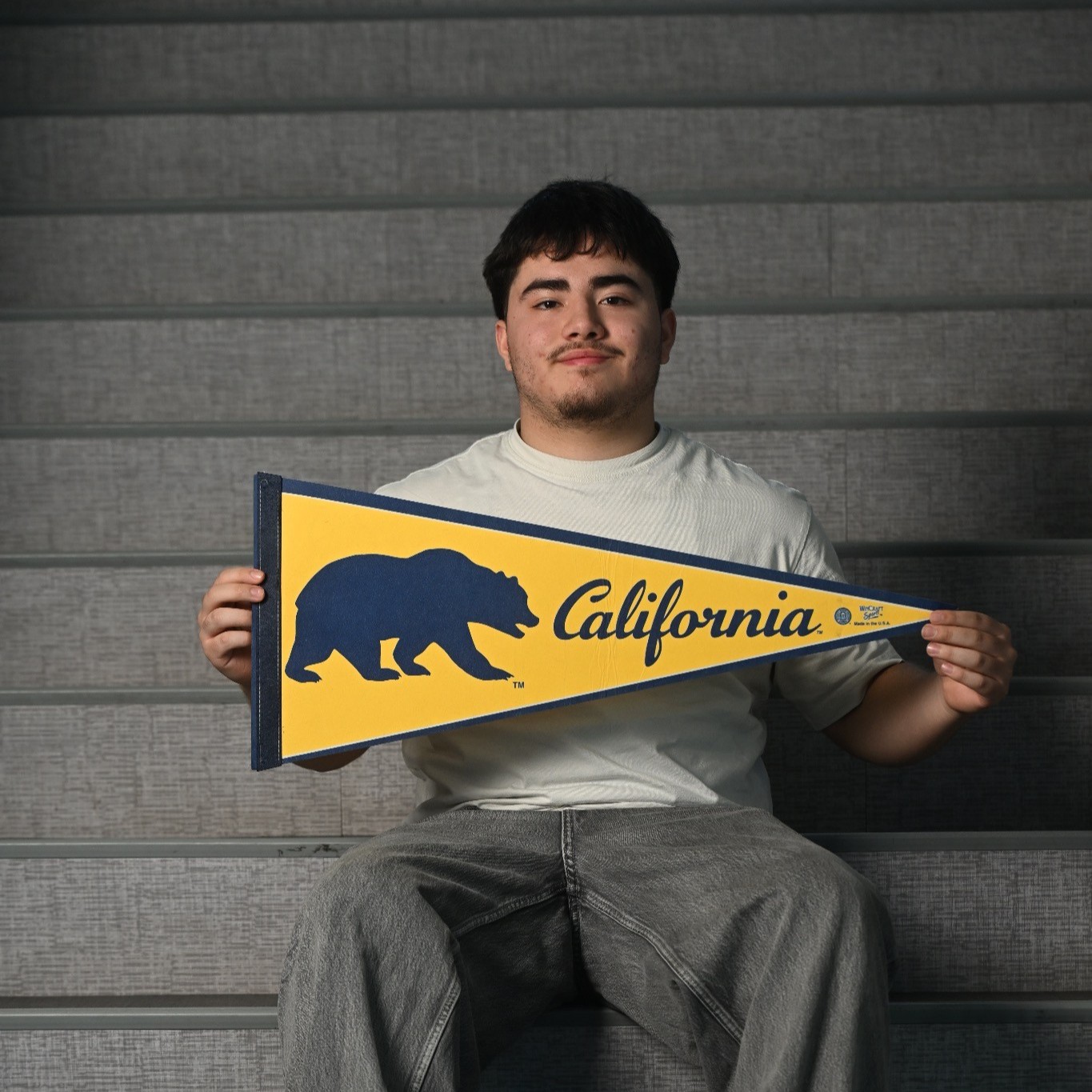 Young man sitting on stadium bleachers, holding a yellow California pennant with a blue bear logo.
