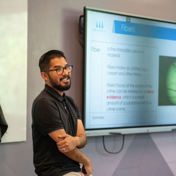 Man with glasses and beard in a black polo stands beside a presentation screen showing a slide about Fibers.