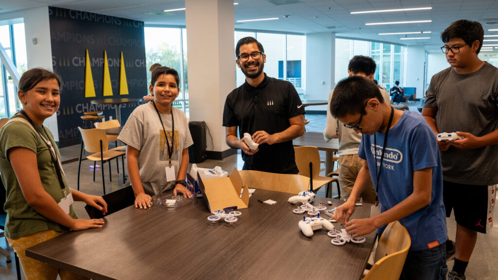 Group of children and a smiling facilitator around a table, assembling white drone controllers in a bright room.