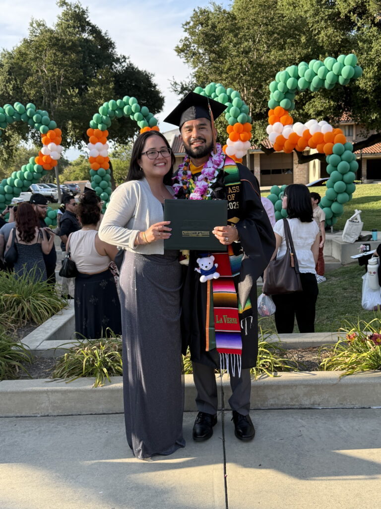 Graduate in cap and gown with colorful lei, holding a diploma folder and posing with a smiling woman outdoors, decorative balloon arches in the background.