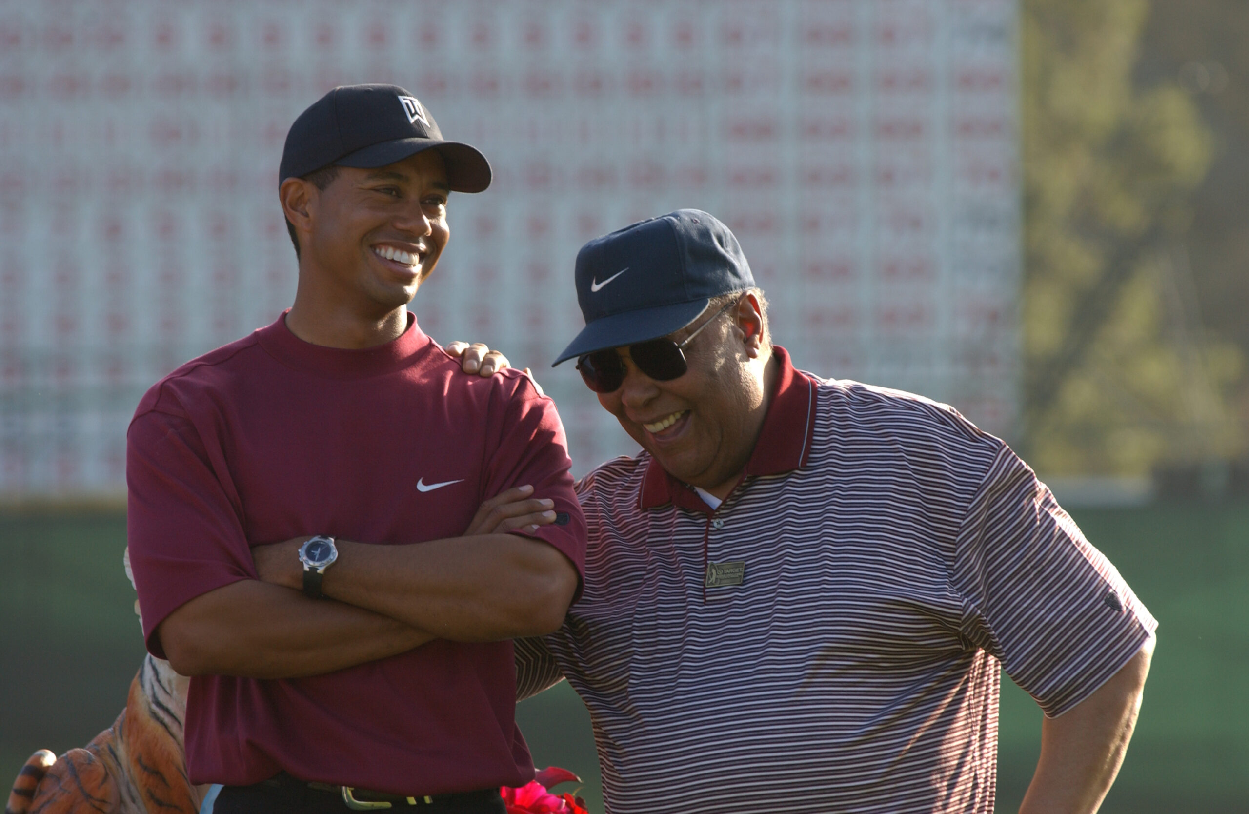 Two men on a golf course laughing and posing together, one in a maroon shirt and cap, the other in a striped polo with sunglasses.
