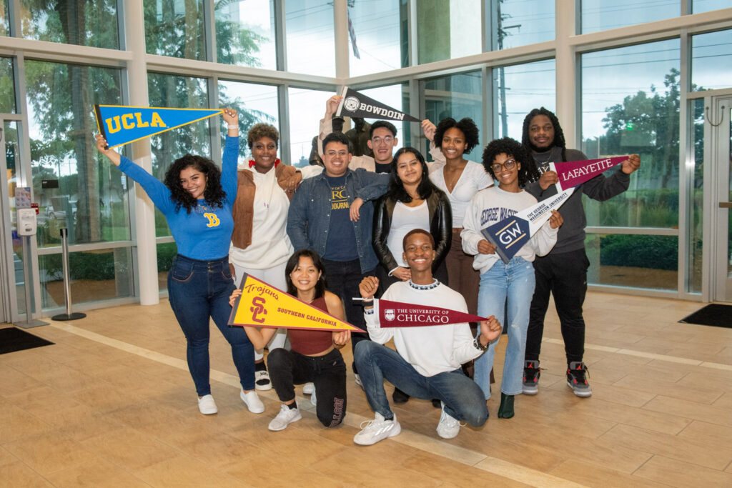 Group of diverse students posing in a campus lobby, each holding a colorful university pennant (UCLA, Lafayette, Chicago, etc.).