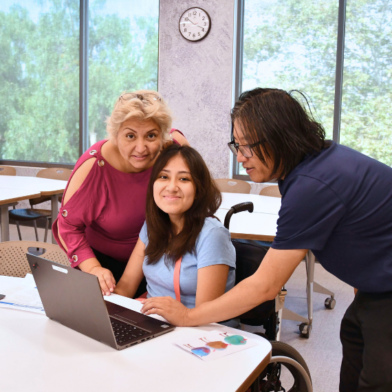 Young woman in a wheelchair works on a laptop as two adults lean in to help in a bright classroom setting.