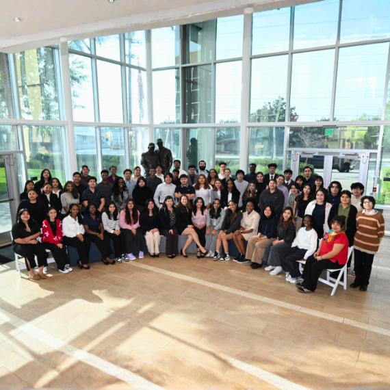 Large diverse group of people posing for a group photo in a sunlit glass-walled atrium.