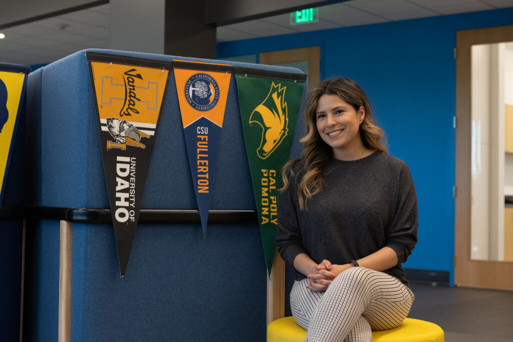 Smiling woman seated on a yellow stool beside a blue wall with colorful university banners hanging from a divider.