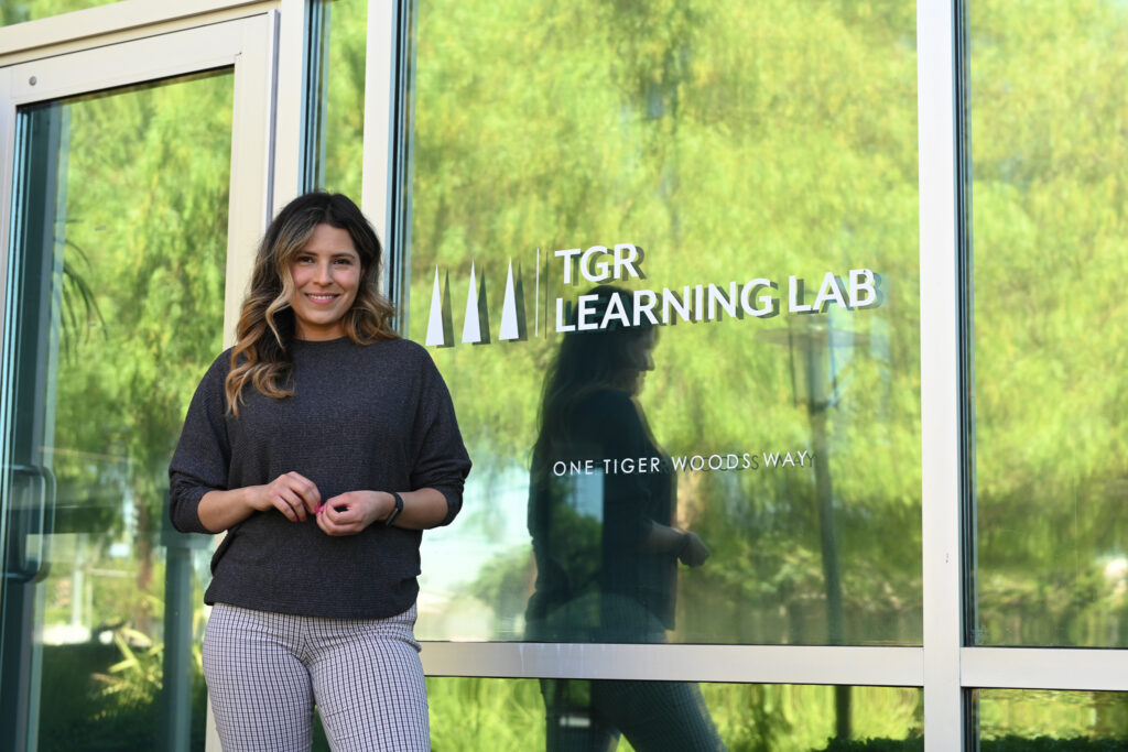 Woman stands beside a glass wall with the TGR Learning Lab logo and greenery reflected in the window, suggesting an outside entrance area.