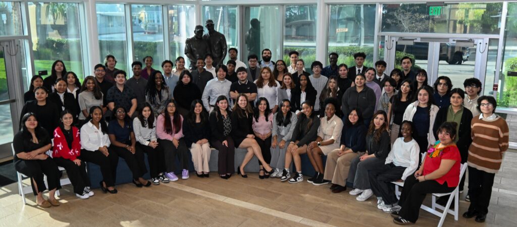 Large group of diverse students posing together in a bright glass-walled lobby with a statue in the background.