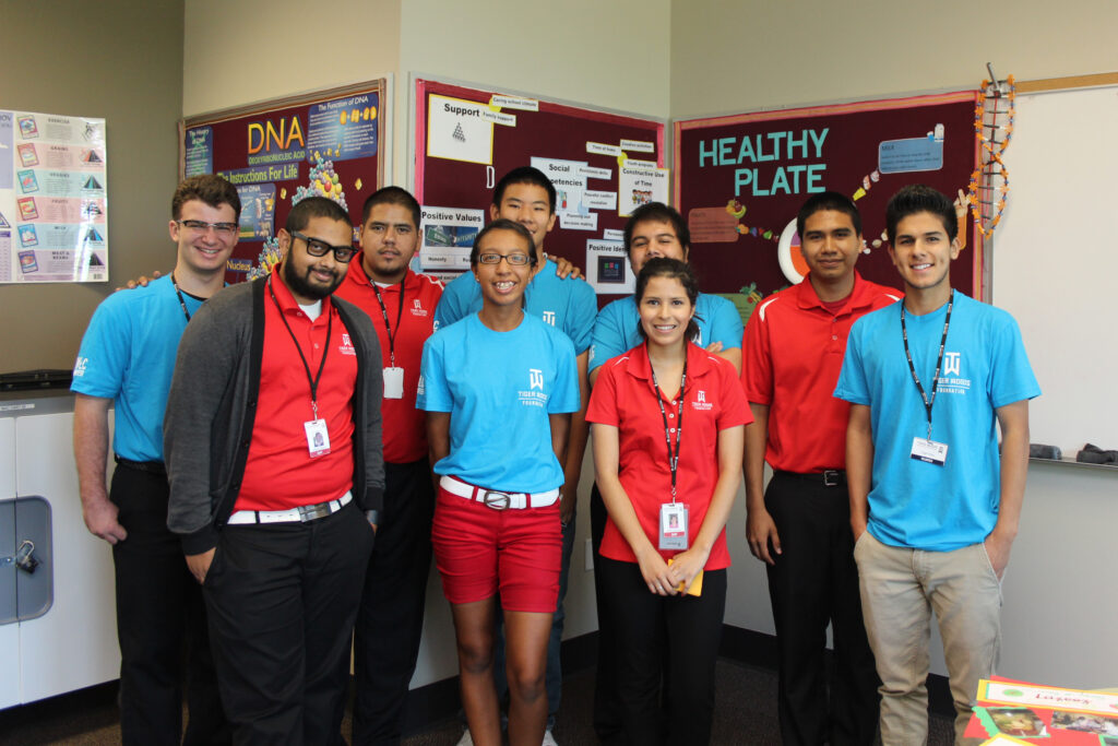 Group of students in red and blue shirts with name badges posing together in a school hallway with science bulletin boards behind them.