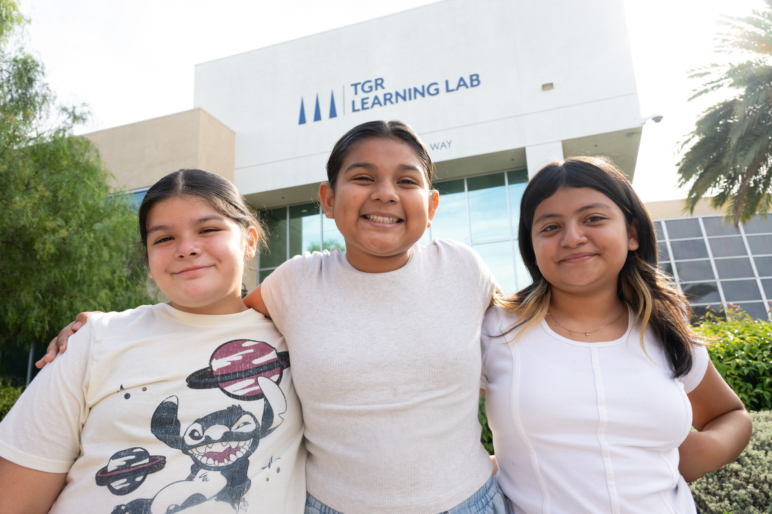 Three smiling girls with their arms around each other stand in front of the TGR Learning Lab building on a sunny day.