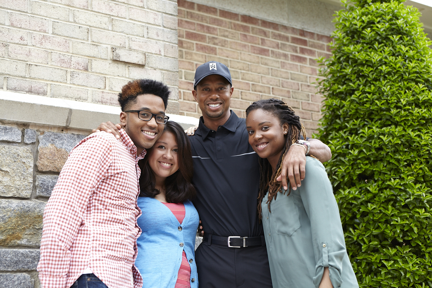 Four friends pose with arms around each other, smiling outside against a brick wall and green hedge.