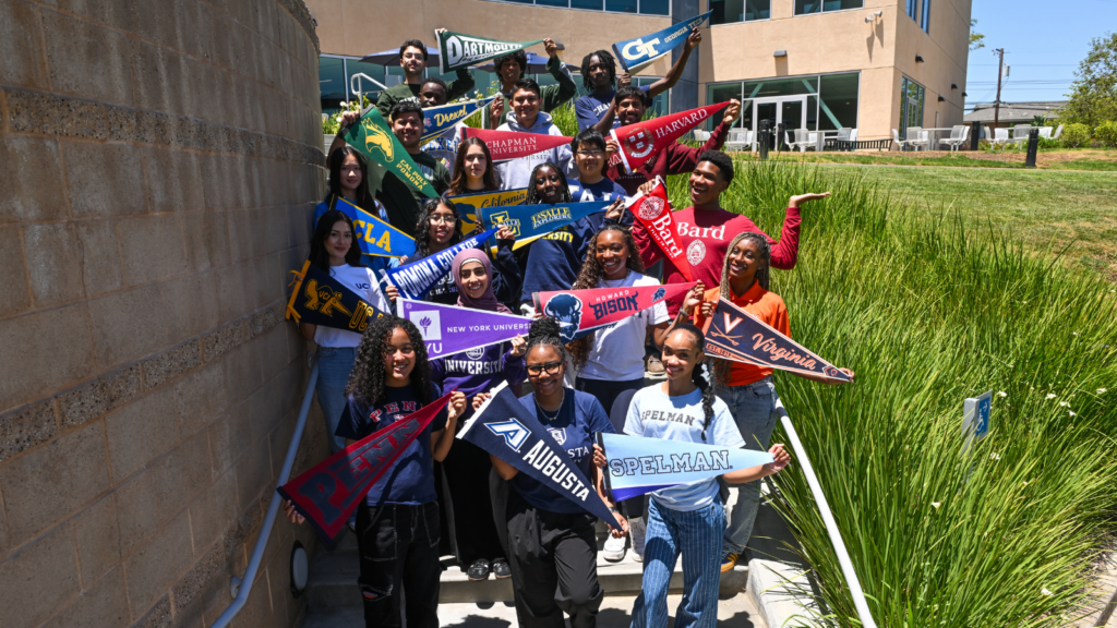 Group of diverse students on a sunny campus stairway, holding colorful college pennants and smiling.