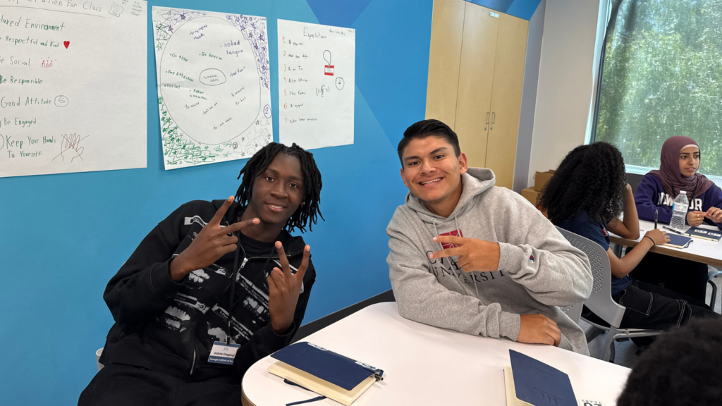 Two students pose and smile at a table in a bright blue classroom, flashing peace signs toward the camera.