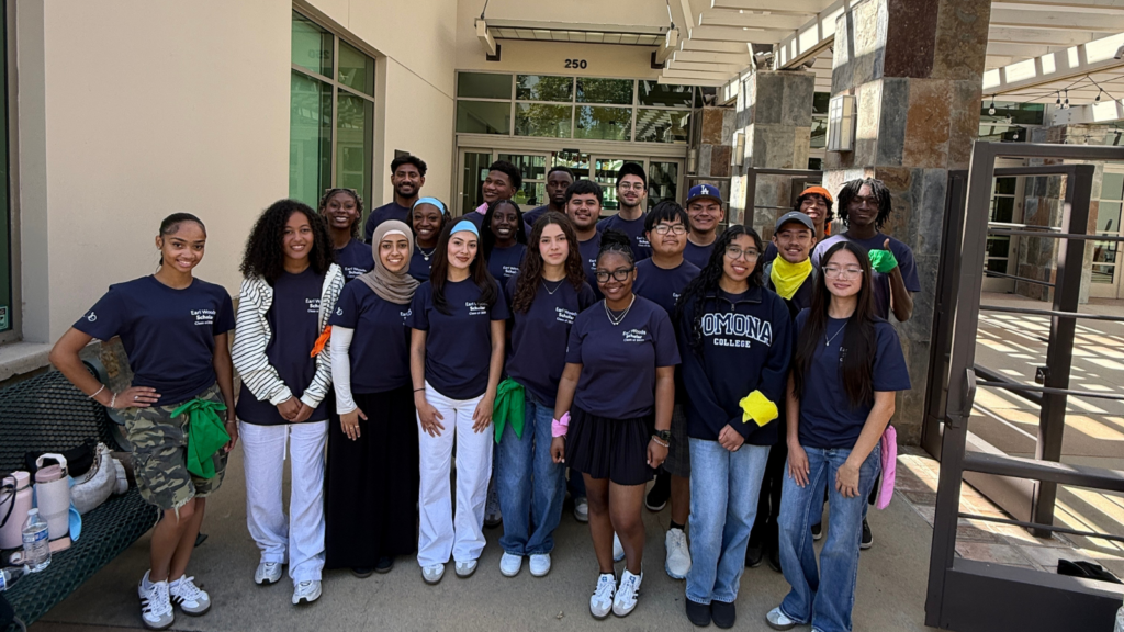 Group of diverse students posing together outdoors in front of a campus building, wearing matching navy T-shirts and casual pants/jeans.