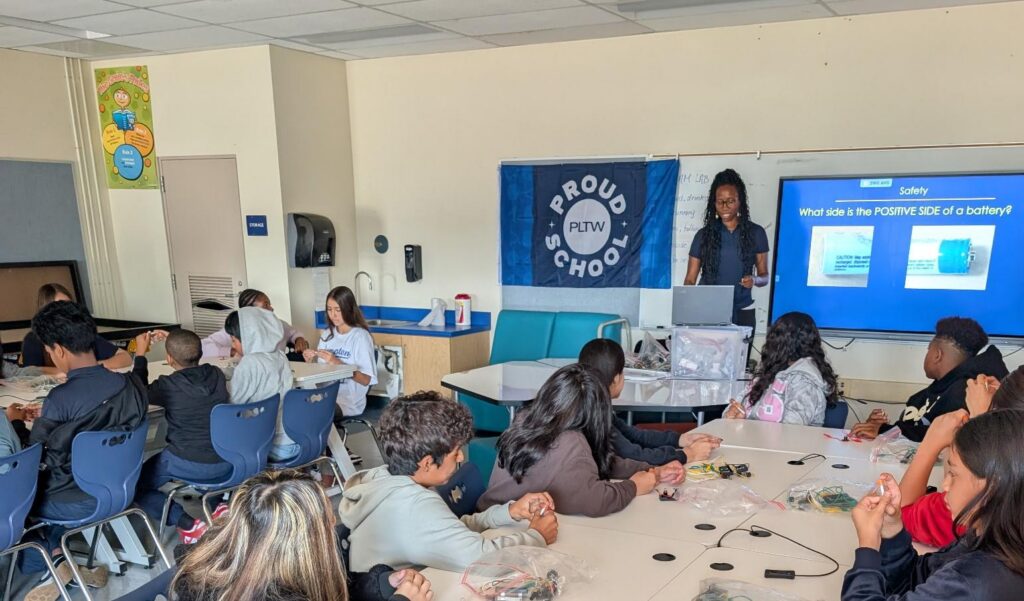 Teacher presents a battery-safety lesson to a classroom of students at lab tables, with a PLTW Proud School banner in the background.