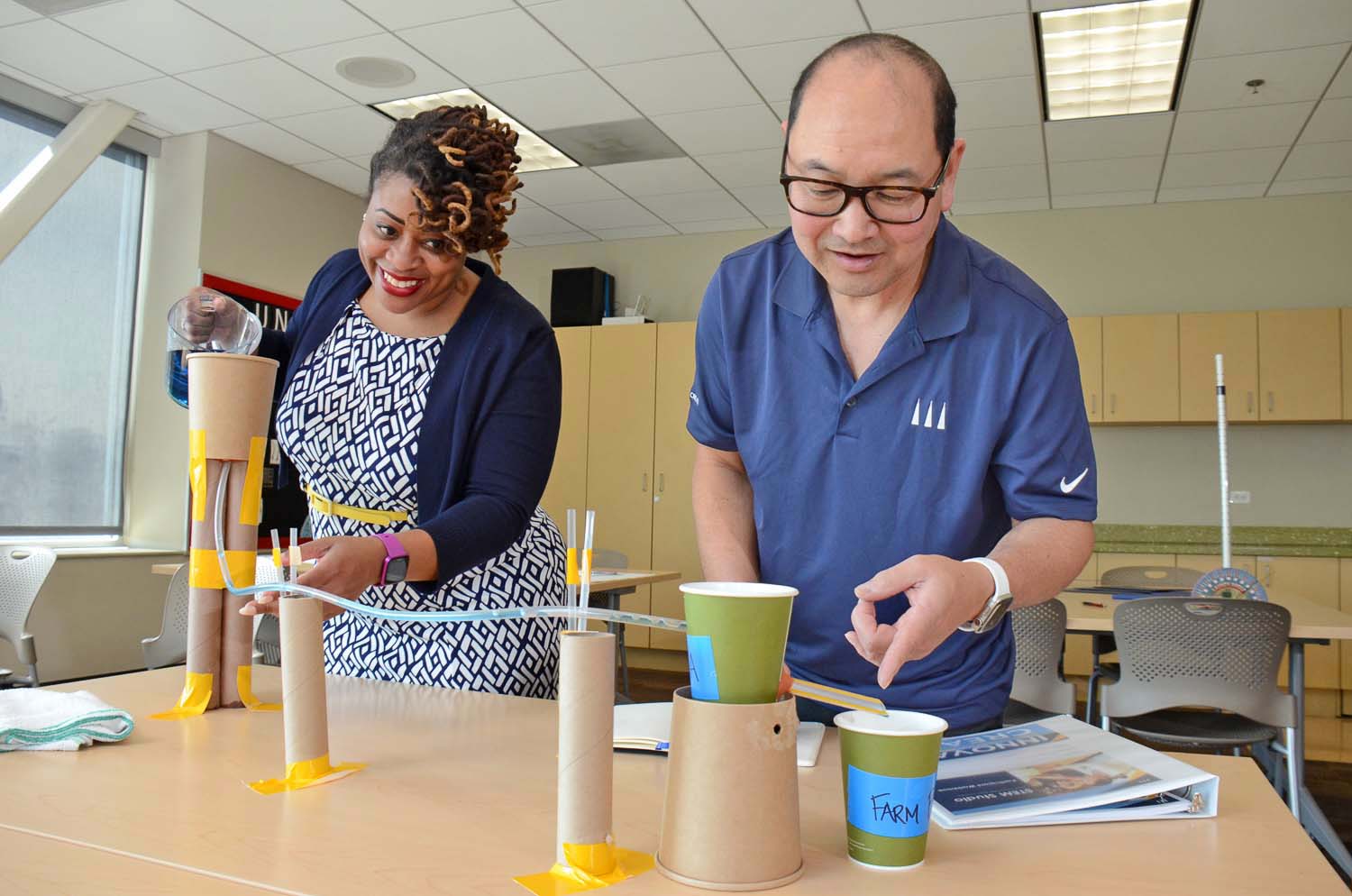 Two adults in a bright classroom build a cardboard-tube project, taping tubes together with string and tape. cups and a notebook sit on the table nearby.