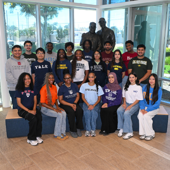 Diverse group of students pose for a class photo in a modern glass-walled atrium, many wearing university T-shirts and smiling.