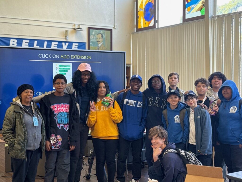 Group of students and adults posing together in a classroom library area with a blue banner reading BELIEVE in the background.