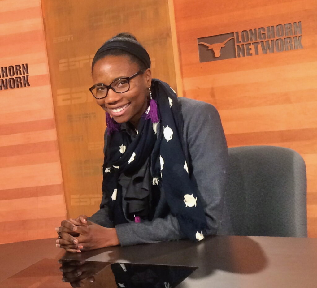 Smiling woman with glasses sits at a desk in a Longhorn Network studio, wearing a dark scarf and gray blazer.