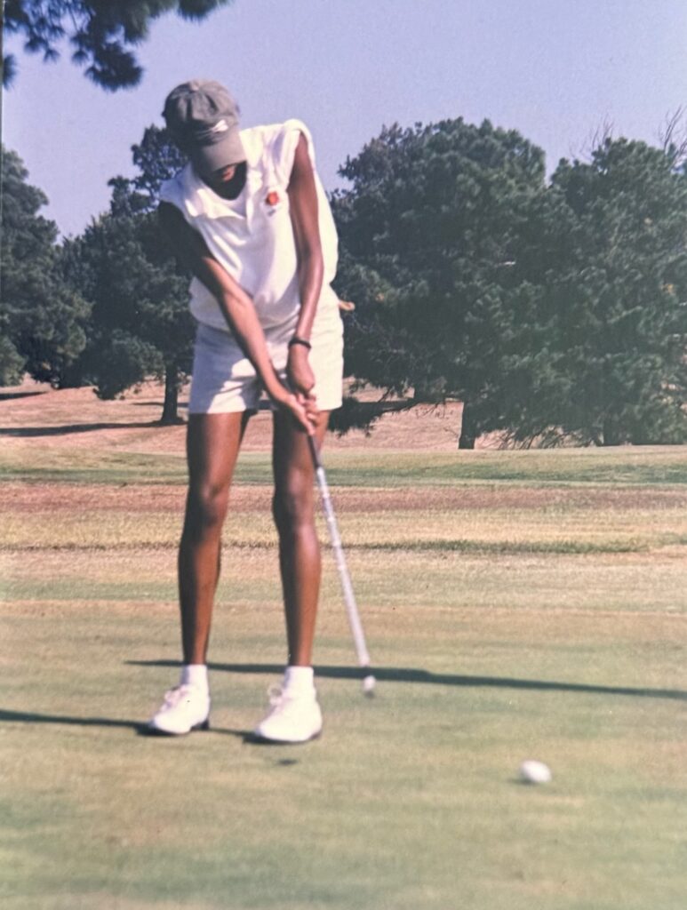 Golfer in a white sleeveless shirt and cap preparing to putt on a sunlit golf course.