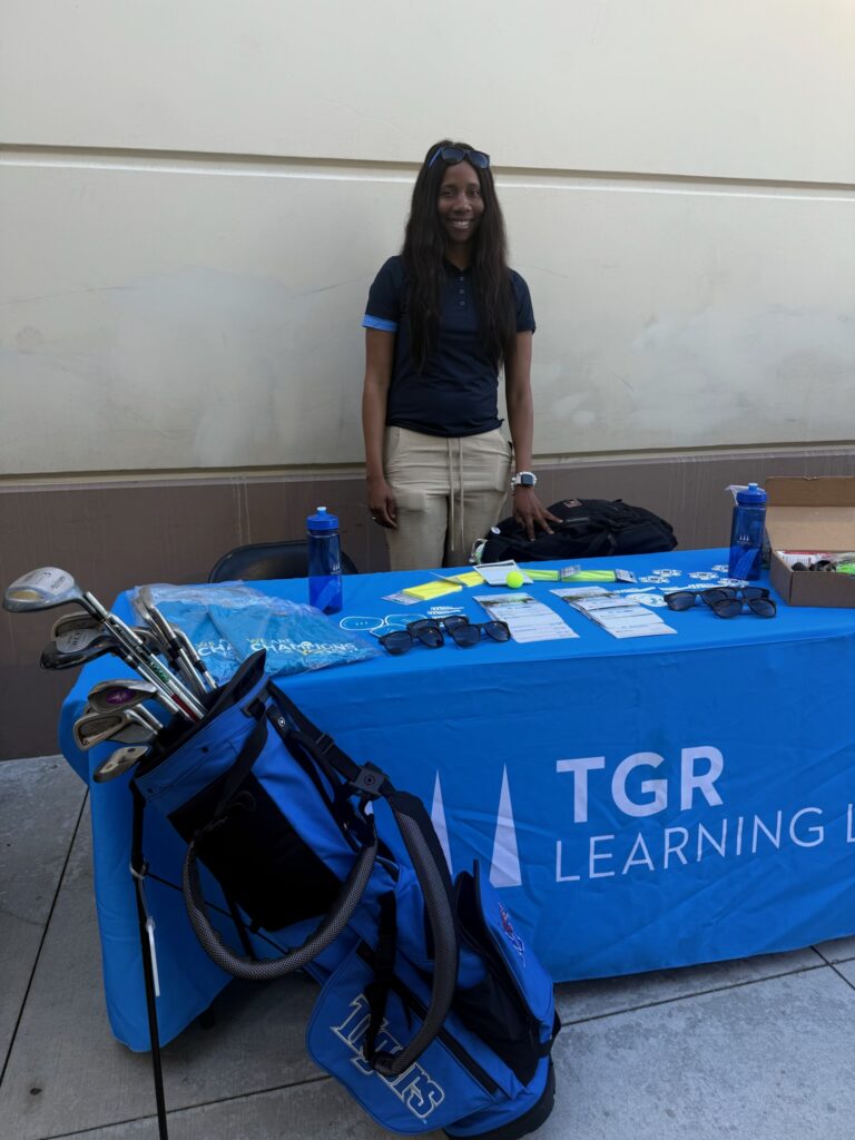 Young woman with long dark hair, sunglasses on her head, stands behind a blue promotional table with golf gear and brochures, smiling at the camera.