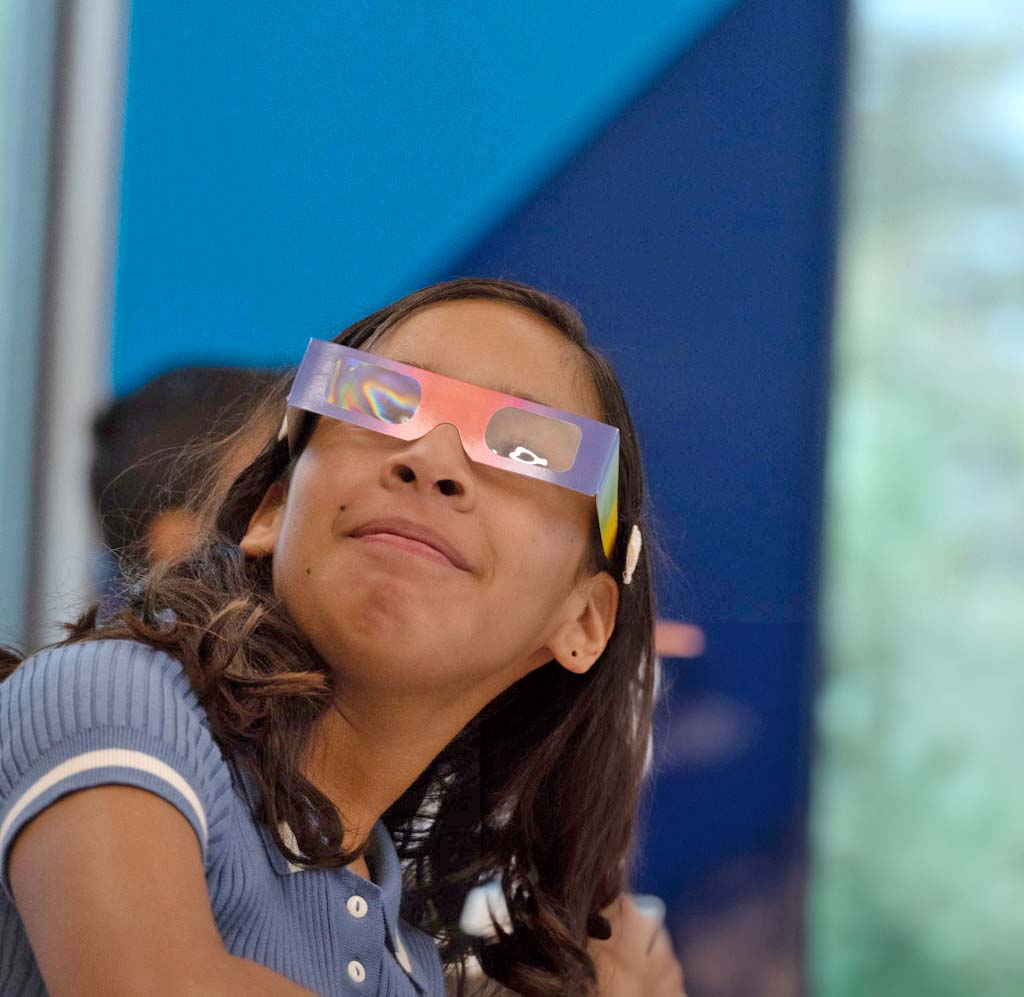 Girl wearing rainbow prism glasses and a blue polo shirt, looking upward with a gentle smile in a bright indoor space.