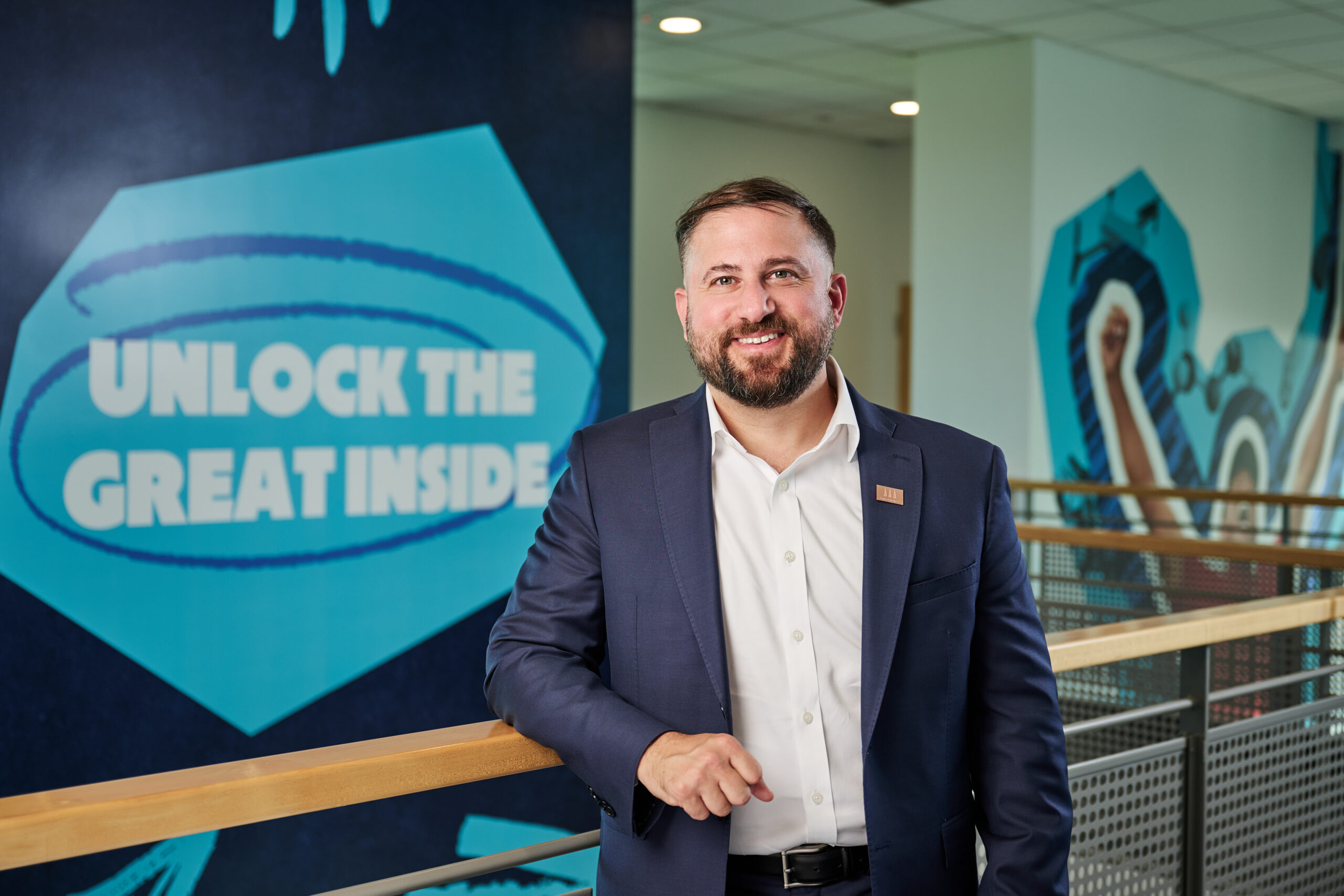Smiling man in a navy suit stands at a wooden railing in a hallway with a blue mural that reads 'UNLOCK THE GREAT INSIDE'.