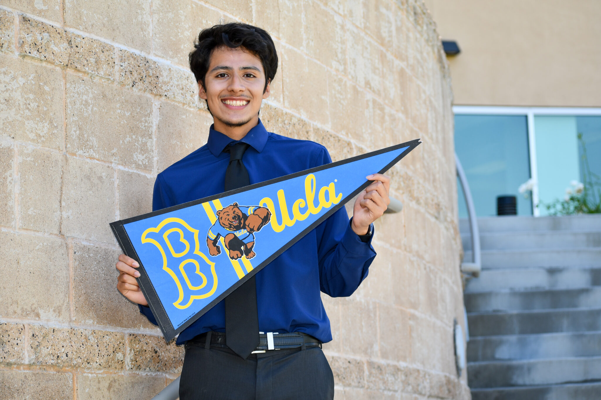 Smiling young man in a blue shirt and black tie holding a blue pennant banner with a bear mascot, standing against a stone wall.