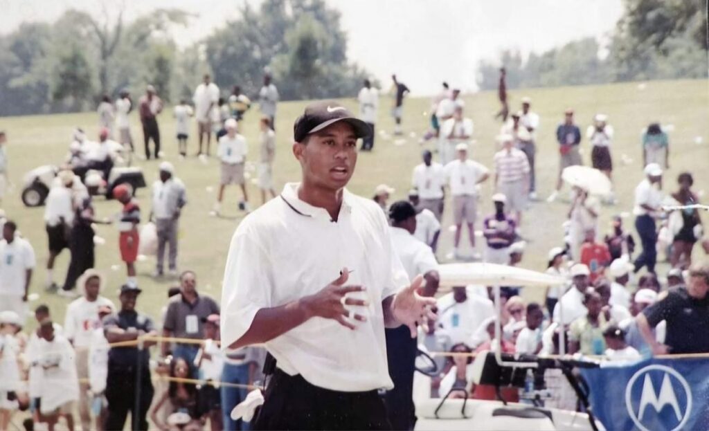 Man in a white polo and black cap speaks to a large outdoor crowd on a grassy field, gesturing with his hands.