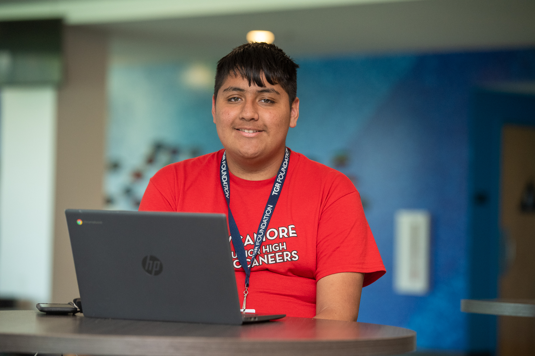 Young man in a red shirt sits at a desk with an open laptop and a TGR Foundation lanyard, smiling at the camera in an indoor setting.
