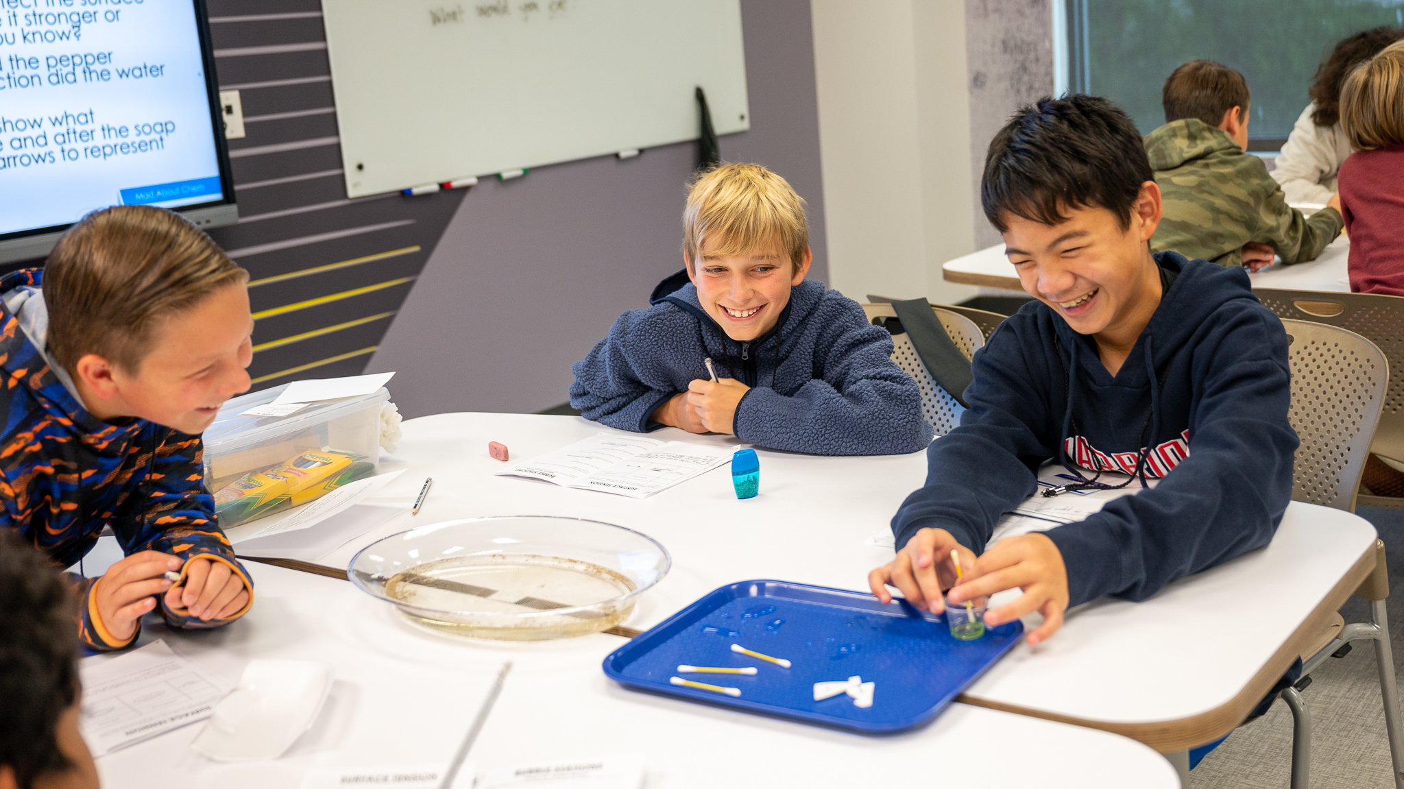 Three boys sit at a white classroom table, smiling as they work on a hands-on science activity with papers and pencils nearby.