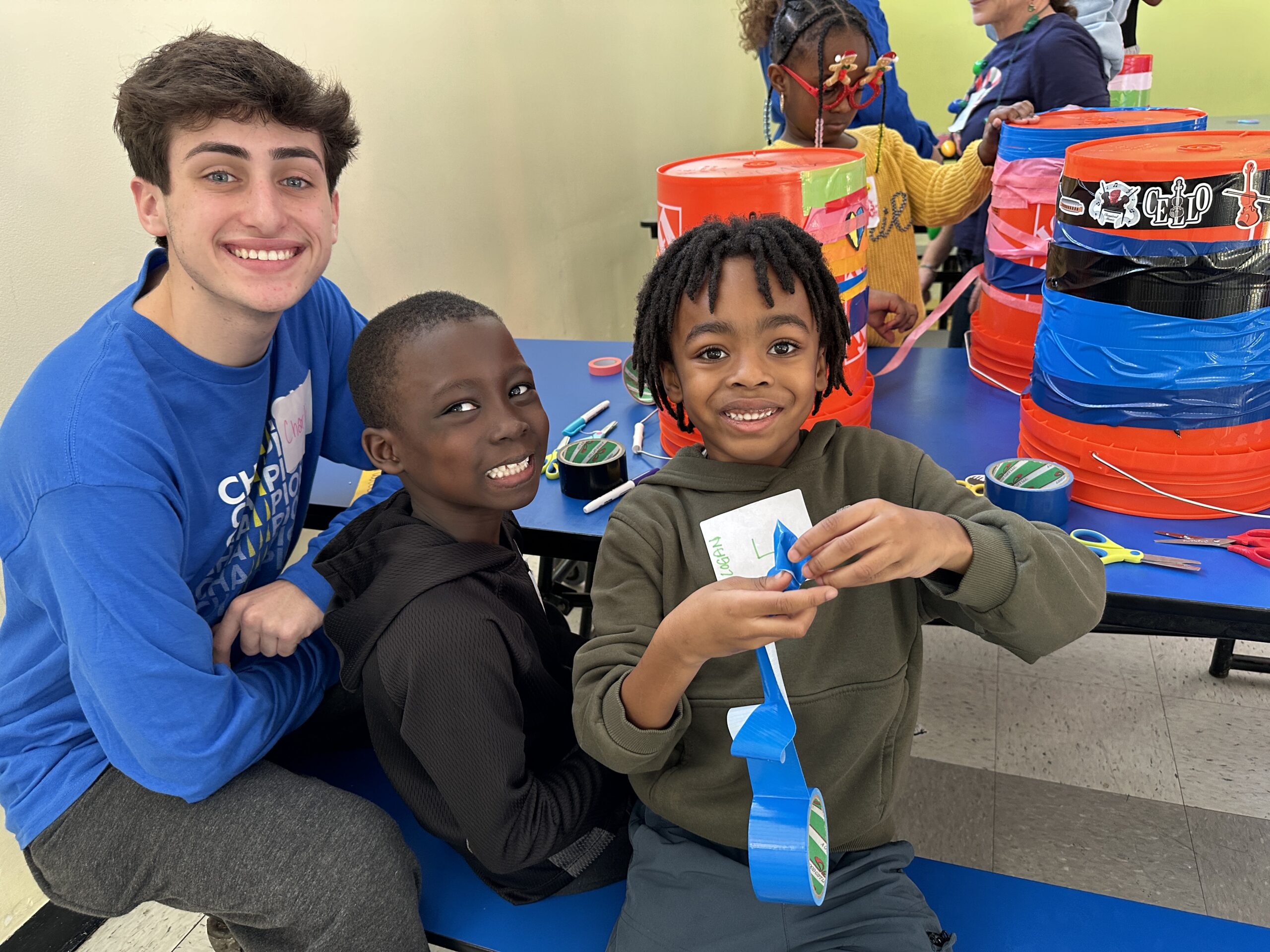 Three kids and a teen sit at a blue table crafting with colorful tape and craft supplies, smiling at the camera.