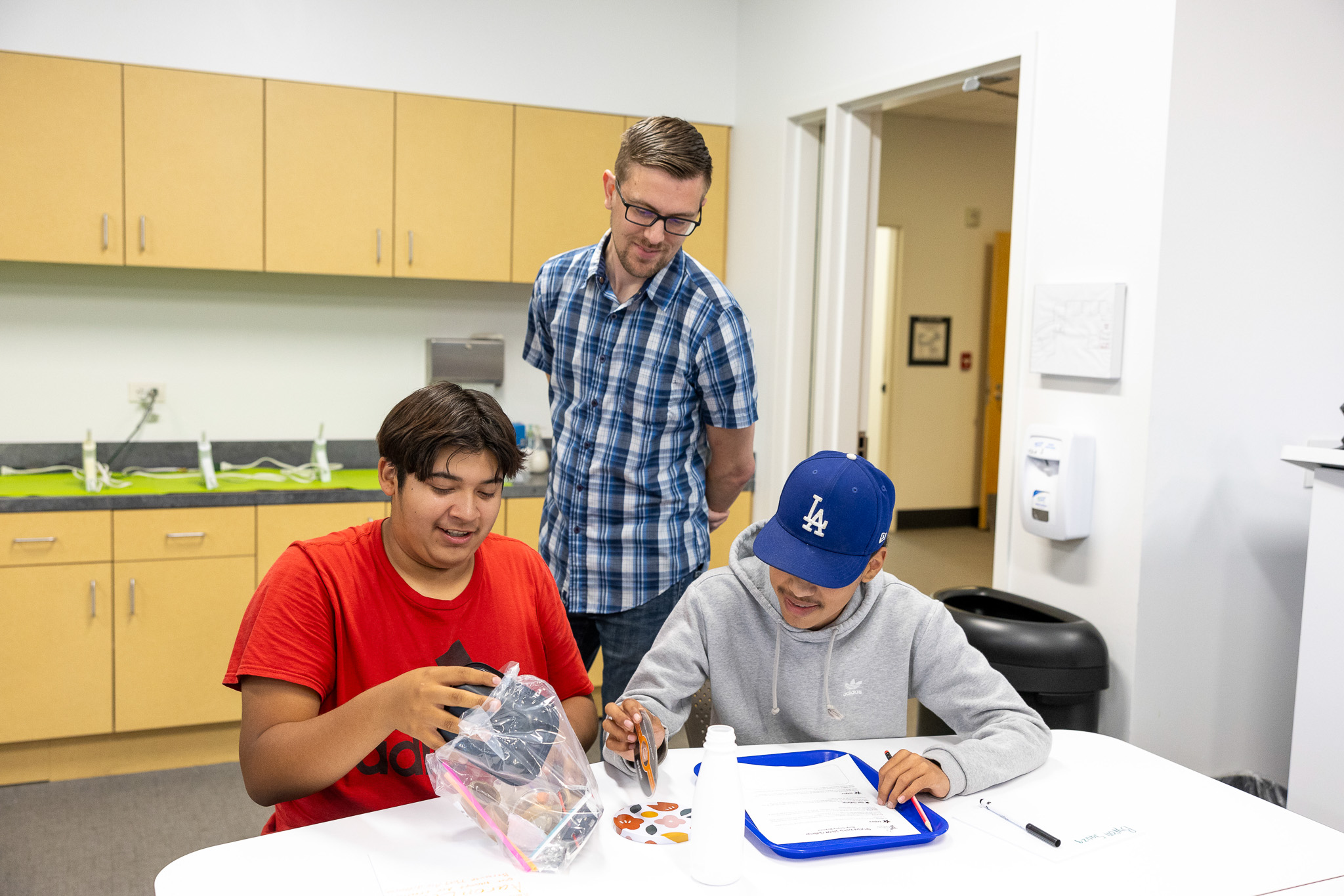 Three students sit at a white table making crafts, while one leans over to help in a bright classroom with cabinets in the background.