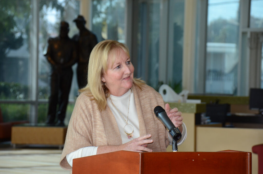 Woman with blonde hair and a beige cardigan speaking into a handheld microphone at a wooden podium in a bright lobby.