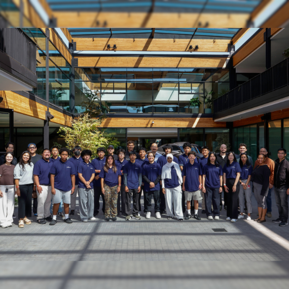 Group of students and faculty posing outside a modern glass-and-wood campus building for a class photo in a courtyard.