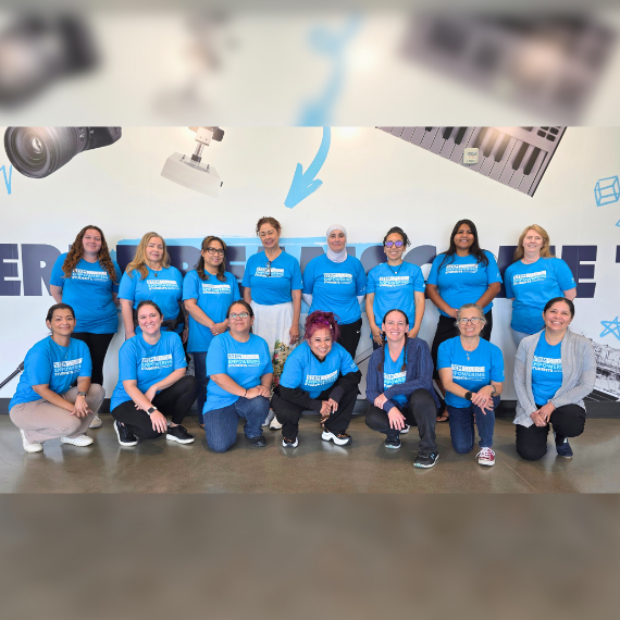 Group of around 15 diverse women in matching blue T‑shirts posing for a team photo in a bright, modern room.