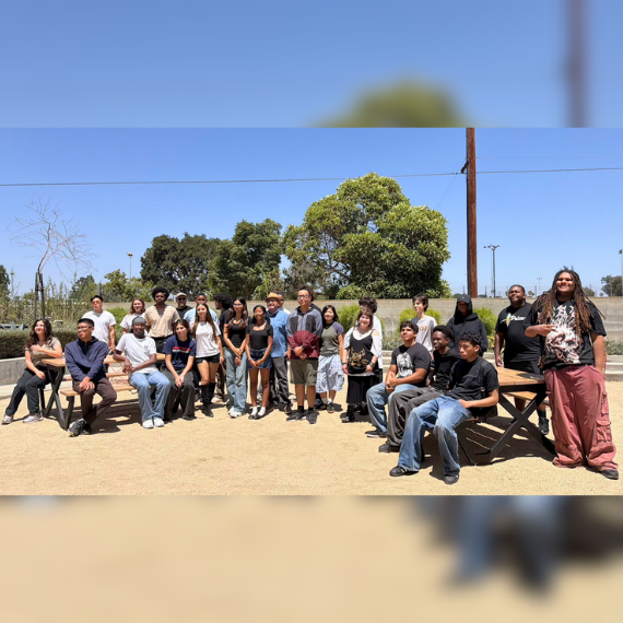 Large diverse group of about 30 people posing for a photo in a sunny outdoor park, with trees and wooden picnic tables in the background.