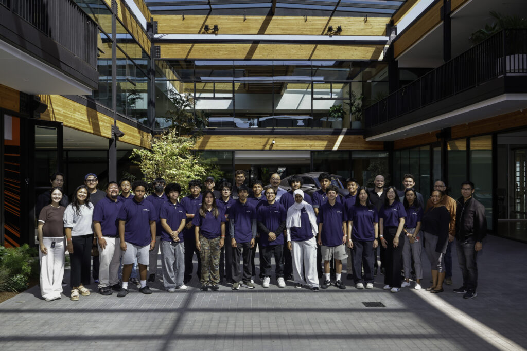 Diverse group of people standing in a modern glass-and-wood courtyard, many in navy polo shirts, posing for a group photo.