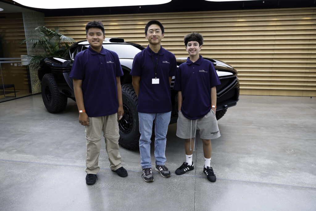 Three young men in navy polo shirts pose in front of a black off-road vehicle inside a modern showroom.