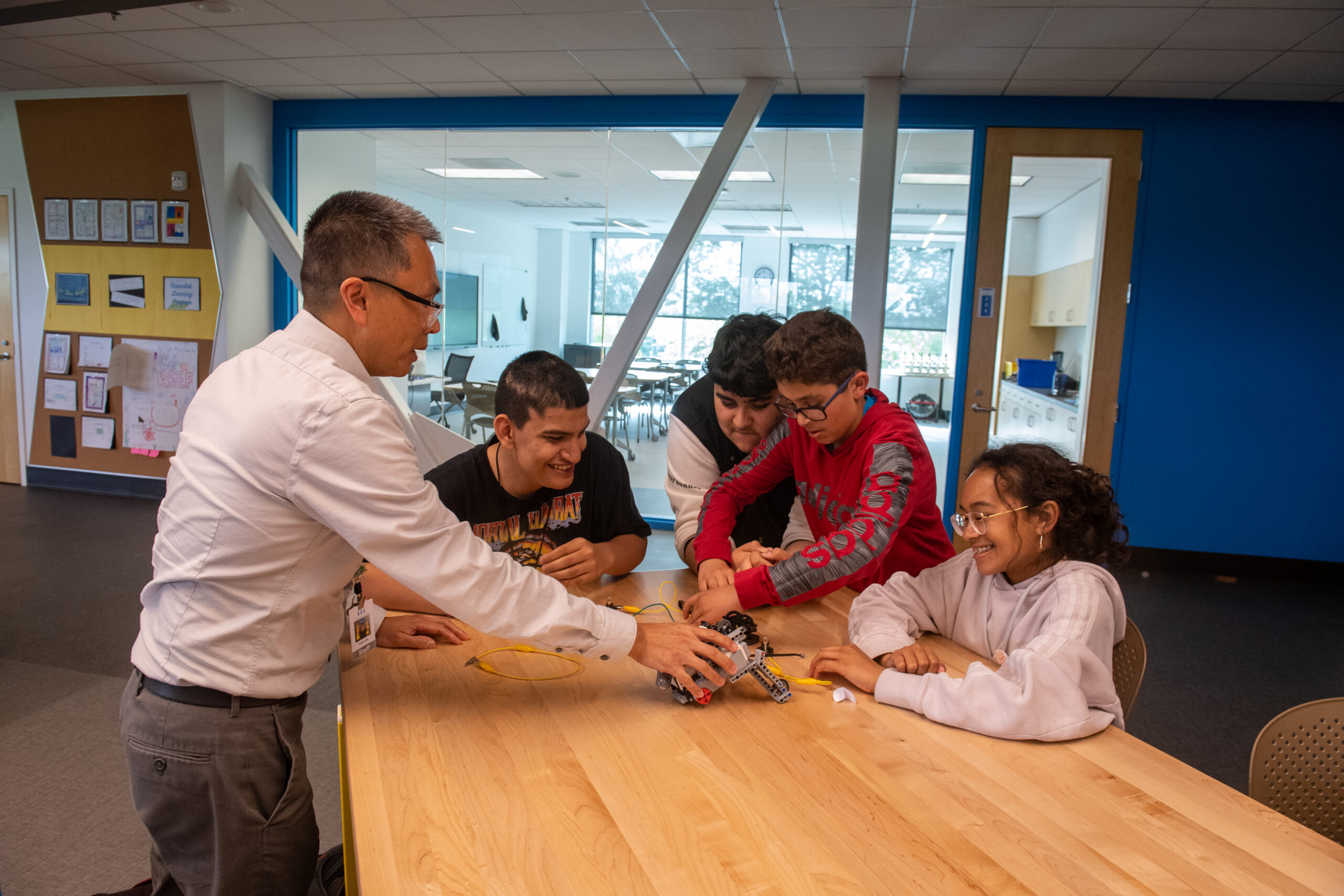 Teacher guiding students as they assemble a robotics kit at a wooden table in a bright classroom.
