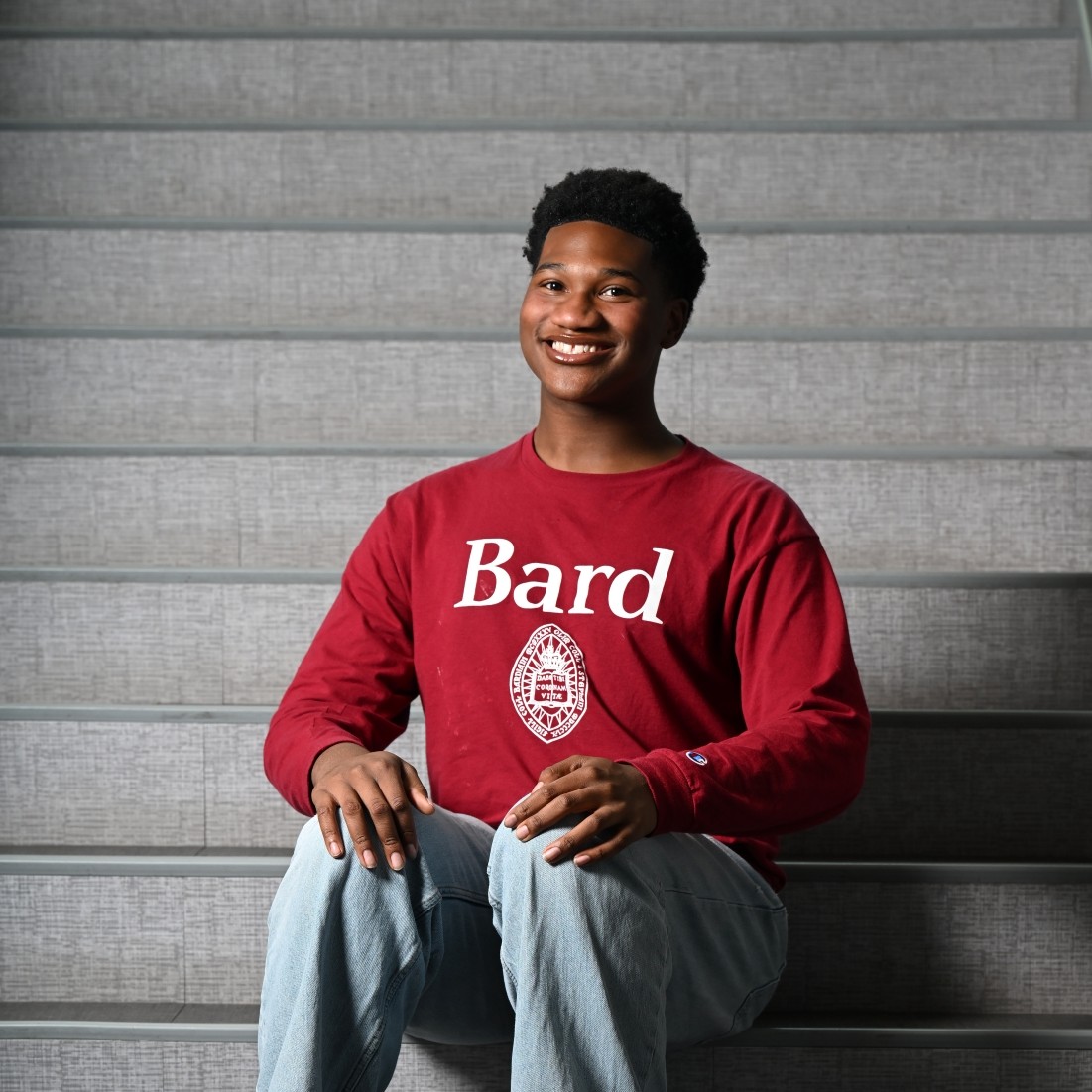 Smiling young man sitting on gray stairs, wearing a red Bard sweatshirt.