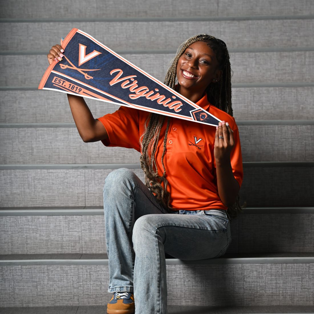Smiling woman in an orange Virginia shirt sits on gray stadium steps, holding a navy and orange Virginia pennant.