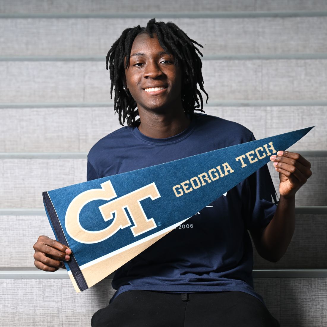 Young man with dreadlocks holds up a Georgia Tech pennant in blue and gold while seated on bleachers.