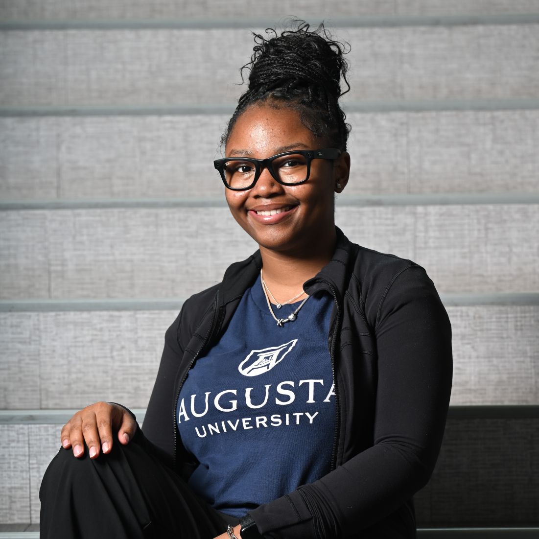 Portrait of a smiling woman with glasses wearing an Augusta University navy T-shirt and black jacket, seated on gray stairs.