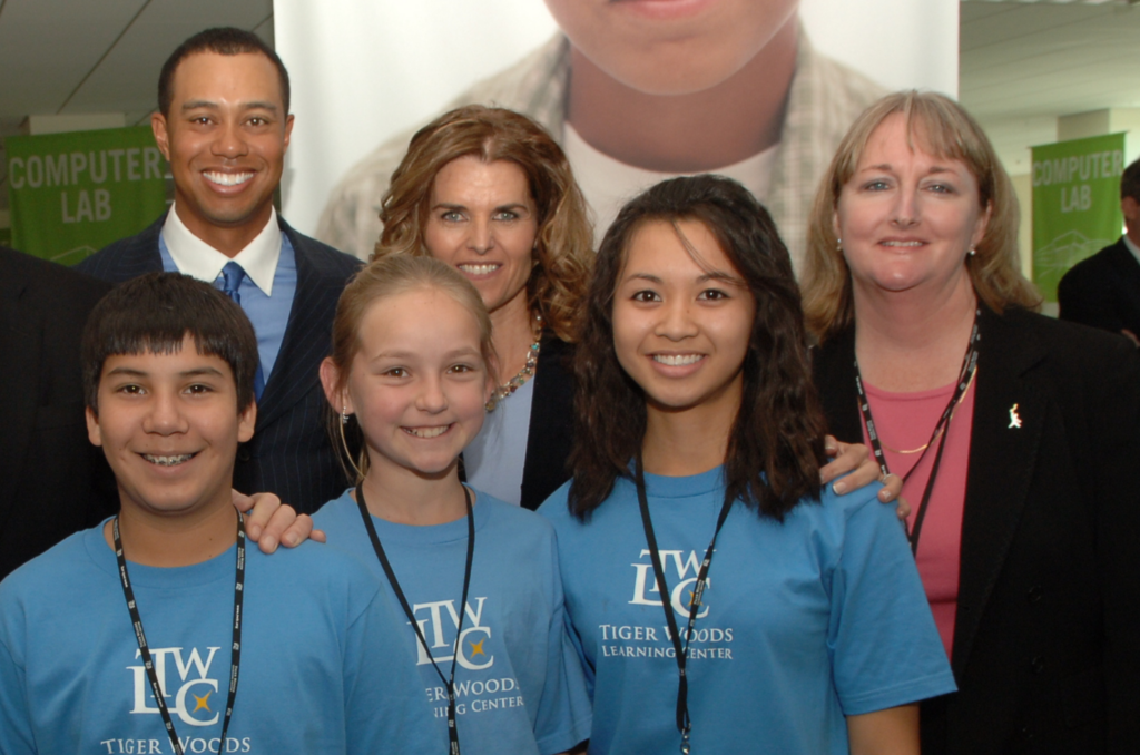 Group of smiling people posing for a photo at a Tiger Woods Learning Center event, indoors with a computer lab sign in the background.