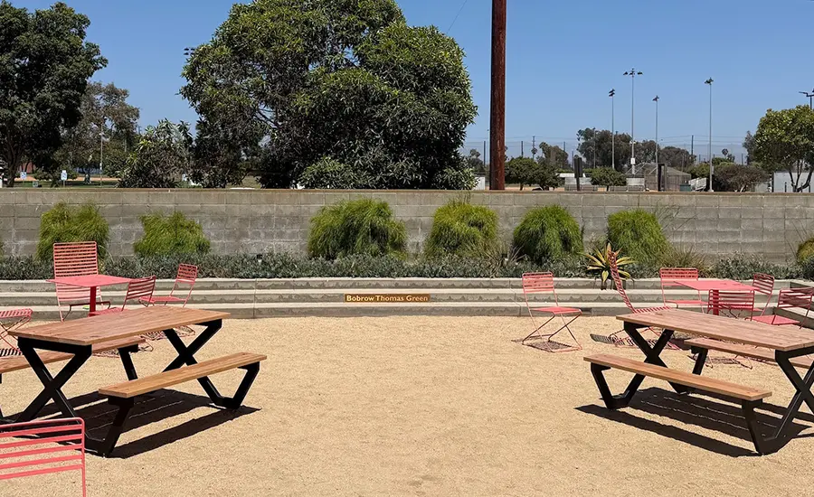 Outdoor park seating area with pink metal chairs and wooden picnic tables on a sandy surface; a concrete retaining wall and greenery in the background, with a small sign on the wall.