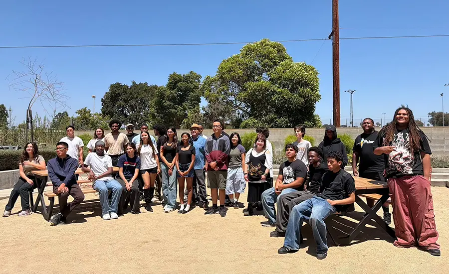 Diverse group of students posing for a group photo in a sunny outdoor park near wooden benches and trees.