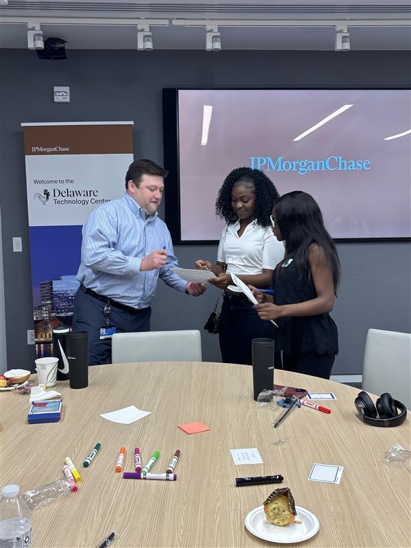 Three professionals stand around a round conference table, exchanging documents near a JP Morgan Chase screen and a Delaware Technology Center banner in a tech center room.