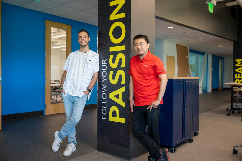 Two smiling men stand beside a tall dark pillar with large yellow lettering in a bright, modern office hallway.