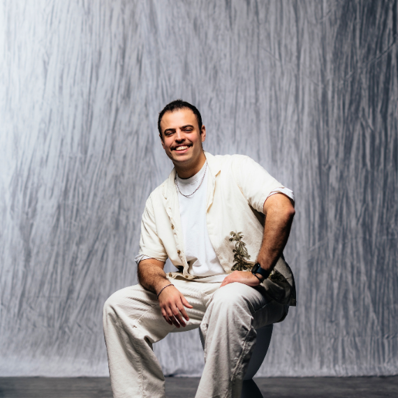 Smiling man in light outfit sits on a stool, small plant tucked into his shirt pocket, posed against a gray textured backdrop.