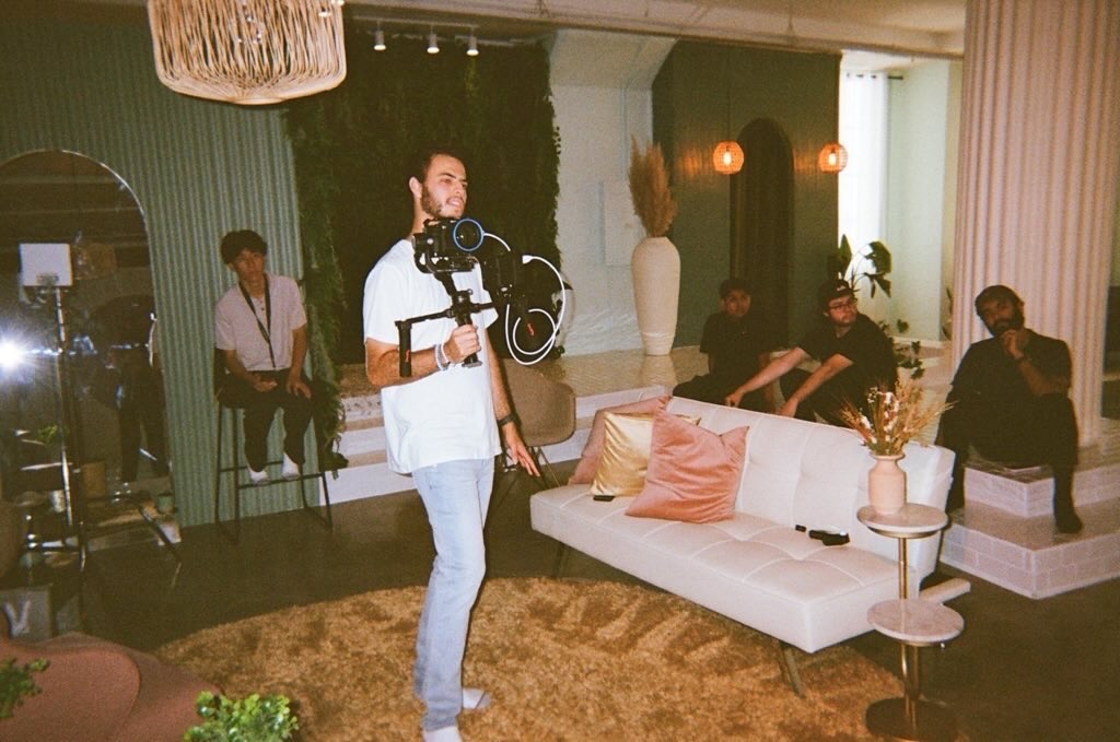 Videographer in a light blue shirt carries a camera rig on a gimbal as a group of people lounge on a white sofa in a stylish living room.",
